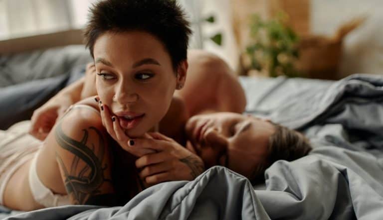 A person with dark hair and tattoos rests in bed next to their partner